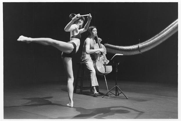 A black and white photograph of two performers in front of a long and curved aluminum duct hung from above. The first performer wearing a bralette and a brief underwear holds a cello bow above their head while extending their right leg straight behind the body. Behind them, the second performer wearing a pair of pants plucks the strings of a cello while looking down at the music stand. The stage has a light color floor and a dark backdrop.