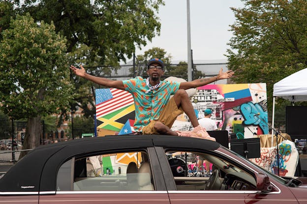 A person sits on the roof of a car with their hands open wide Behind him sits a collage containing various flags and graffiti. 