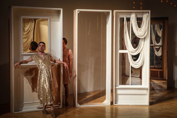 A performer with short hair and a gold sequined dress stands in front of a makeshift window on stage. Another person stands behind the window in-between its border and another makeshift window's with white drapes border.