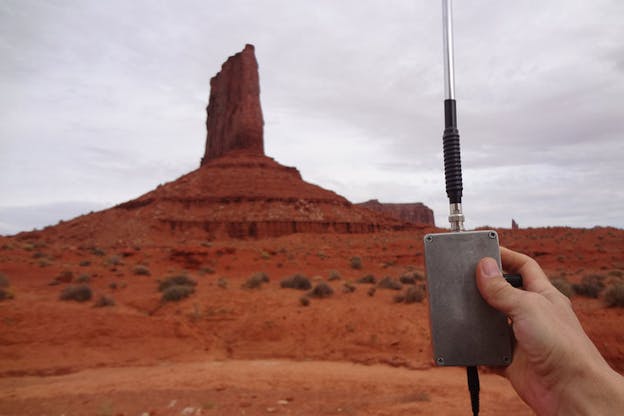 A hand holds out a metal gray low frequency receiver in front of a cinder red desert rock formation.