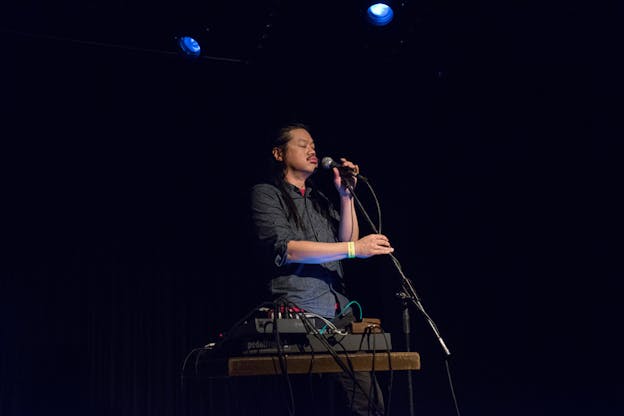 Photograph of Spencer Yeh singing on a microphone behind a desk supporting a pedal board with various cables.