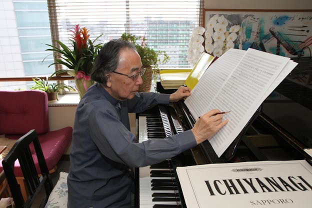 A person in profile dressed in glasses and a gray button up seated at a piano, reads a music sheet situated on the piano. In the front right closest to the viewer a white sheet with the word 