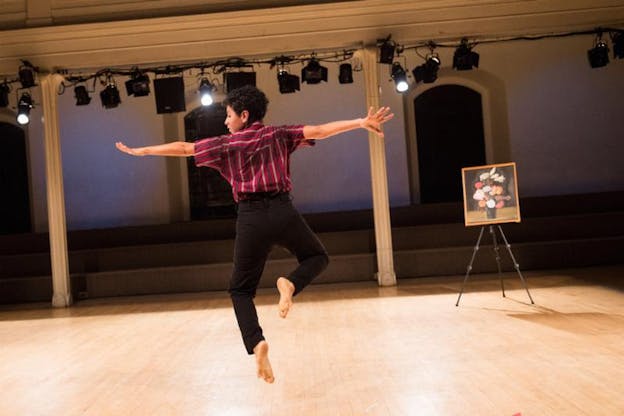 A performer dressed in black pants and burgundy striped shirt with their back to the viewer, jumps in the air with open arms. In front of them on the wooden floor a painting of flowers held by a tripod.