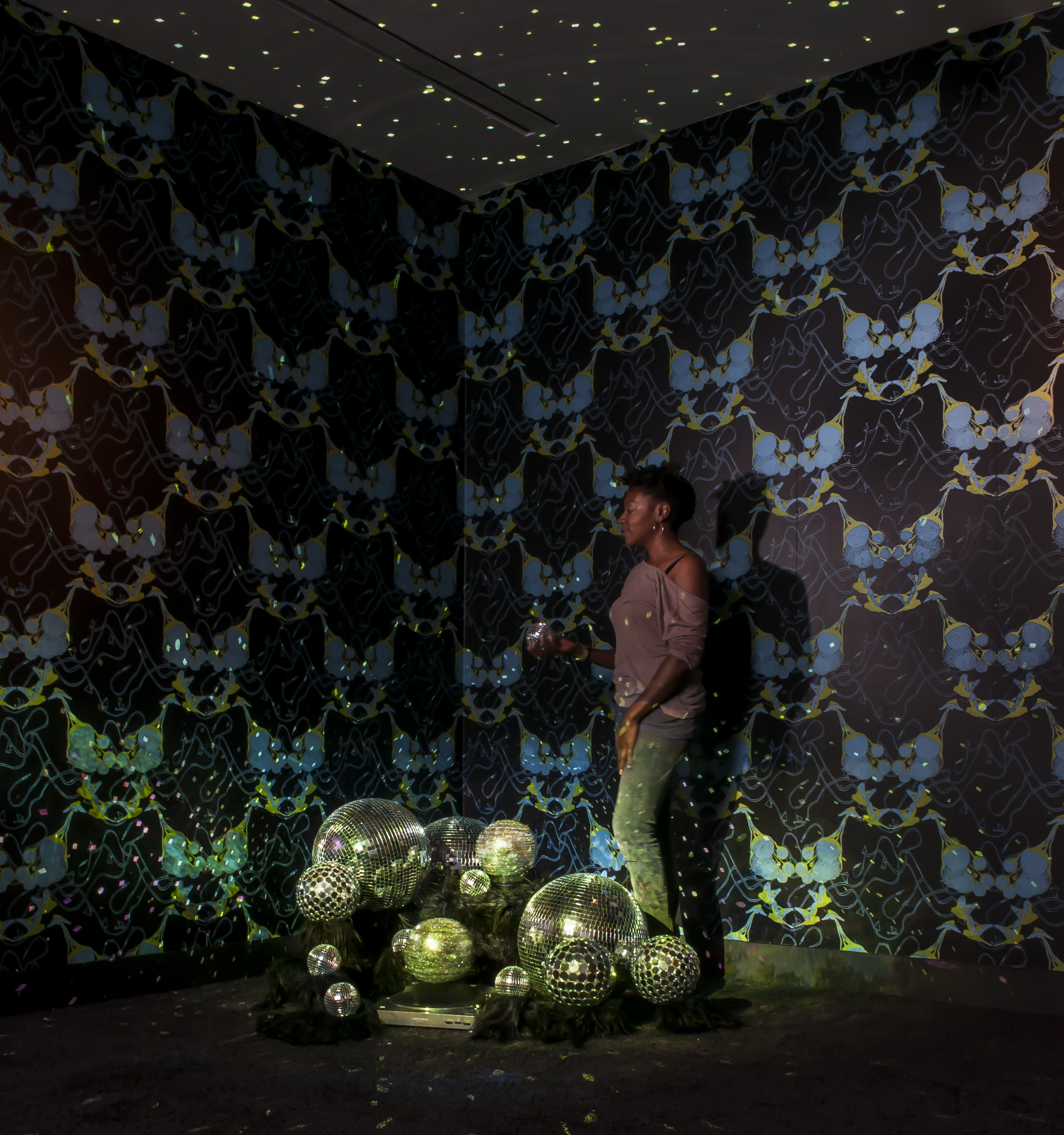 Performer stands in a dimly-lit room speckled with disco ball lights and wallpapered with yellow, dark blue, and pastel blue patterns, holding a tiny disco ball in one hand. On the floor is a pile of disco balls varying in size.