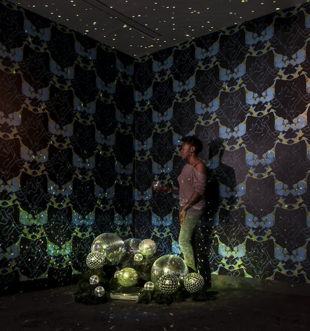 Performer stands in a dimly-lit room speckled with disco ball lights and wallpapered with yellow, dark blue, and pastel blue patterns, holding a tiny disco ball in one hand. On the floor is a pile of disco balls varying in size.