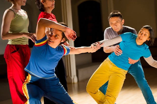 A smiling performer leans outwards with their arm raised above their brow, grasping the wrist of a performer raising their eyebrows expressively while supported by another performer.