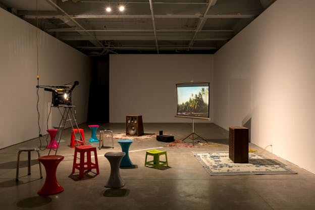 A dimly lit room with multi-colored stools and retro wooden speakers atop carpets beside a projector and screen display of a tropical beach with palm trees.