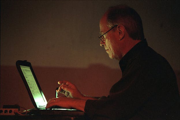 Bischoff sits in a dark space in front of a green illuminated laptop screen, looking intently at the screen and types on its keyboard. He wears a dark shirt and glasses. 