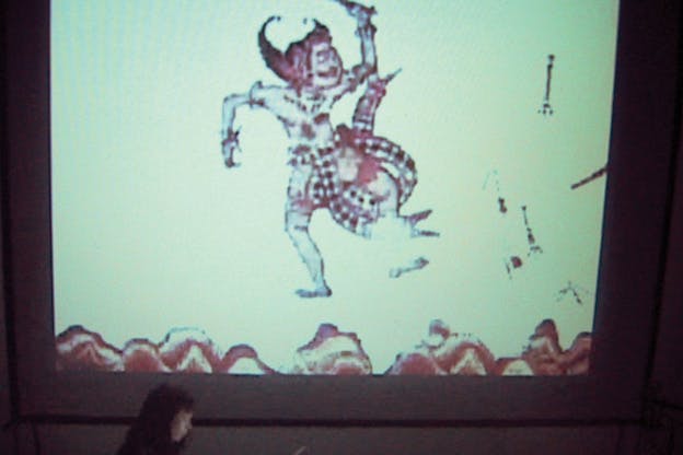 A projection of an illustrated Japanese dancing figure floating on top of mountain ranges in white and terracotta colors. Ikue Mori is situated below the projected screen, showing the side profile of her head.