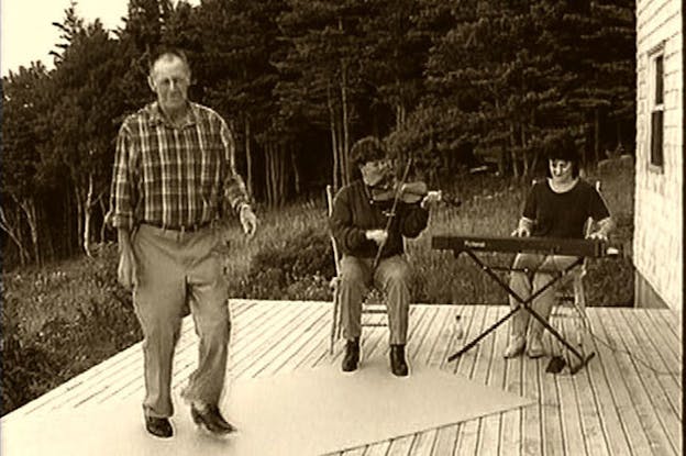 A sepia colored photograph shows a man tapdancing on smooth surface while two sitted women behind him play musical instruments. Behind them trees extend.