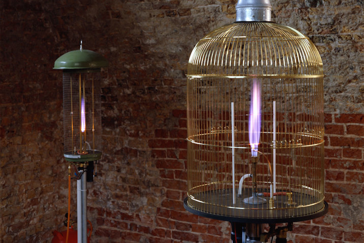 Two bird cages, one in the further front and one in the back, have fires lighted blue inside of them. Next to the birdcage furthest behind a small red tank is situated linked to a cable connected to the bottom of the cage. A brick wall sits behind the two cages. 