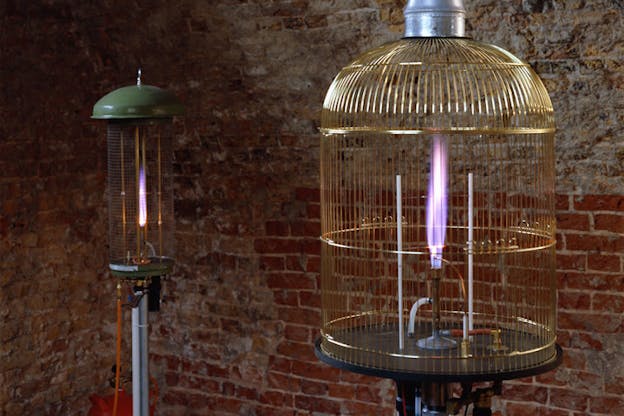 Two bird cages, one in the further front and one in the back, have fires lighted blue inside of them. Next to the birdcage furthest behind a small red tank is situated linked to a cable connected to the bottom of the cage. A brick wall sits behind the two cages. 