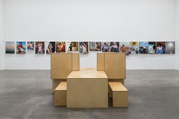 A wooden structure compromised of blocks and benches put together stands in front of a aligned images of people hanging from a horizontal cord on a wall. 