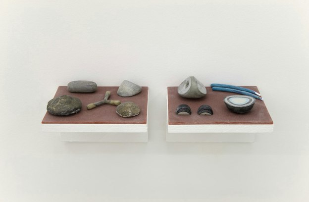 Front view of white and brown painted wooden shelves support objects shaped like a forked twig, geods, rocks, and blue painted sticks. 