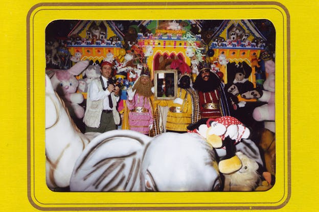 A photograph is surrounded by yellow paper. In the photograph, people in play royal clothing stand in front of a whimsical yellow castle and surrounded by toys. They hold up a mirror. One person on the far left holds up a camera. 