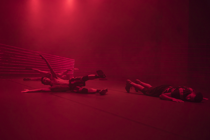 Three performers in a red lighted room lay on the floor. Two of them laying sideways prop their arms and one leg up, while the third lays down on their front.