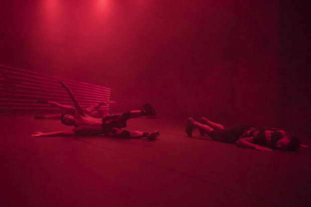 Three performers in a red lighted room lay on the floor. Two of them laying sideways prop their arms and one leg up, while the third lays down on their front.