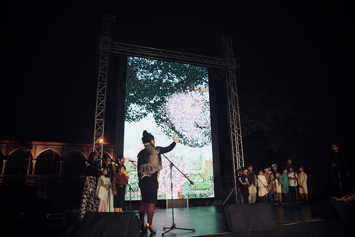 On the stage a children's choir is separated in two groups by a projection of pink and purple swirls in a grassy environment. Closest to the viewer a figure with their back turned, dressed in high heels and a dress directs the choir with their hand.