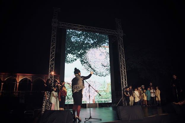 On the stage a children's choir is separated in two groups by a projection of pink and purple swirls in a grassy environment. Closest to the viewer a figure with their back turned, dressed in high heels and a dress directs the choir with their hand.