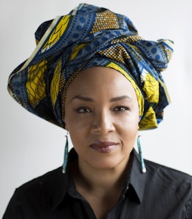 A portrait of LaTasha Diggs wearing a yellow and blue headpiece and a black button up, smiling towards the camera.