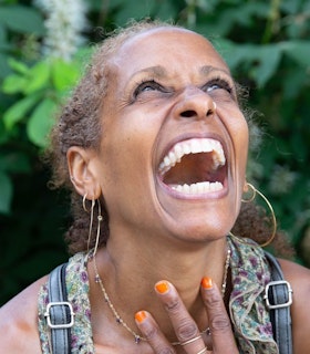 A portrait of nia love in front of a blurred background of green foliage. She looks up, tilting her head back and opening her mouth in an expression of joy. She wears thin gold hoop earrings, a thin gold necklace, a blue floral patterned top, blue overalls, and orange nail polish. 