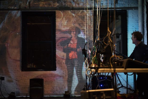Person in right corner observes the images of a full-length person juxtaposed in front of a zoomed-in photograph of the person's face projected onto a white-painted brick wall. 