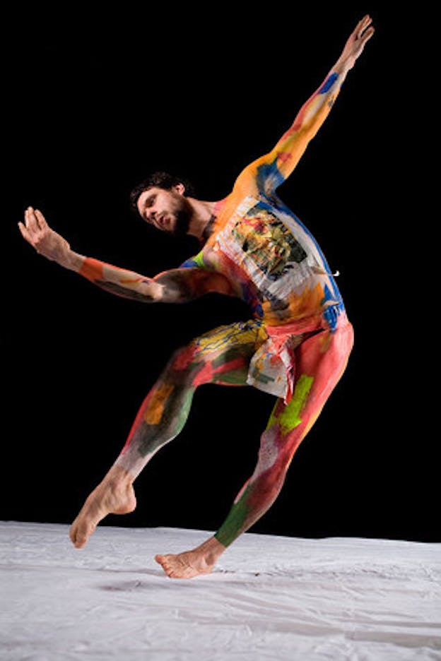 A dancer whose body is painted from neck to ankle in earthy oranges, reds, greens, and blues leans to the side, supported only by the outside edge of their left foot. Their right knee is bent and their right foot is pointed and arched. Theirleft arm is straight and raised behind them and their right arm is bent in front of them. 