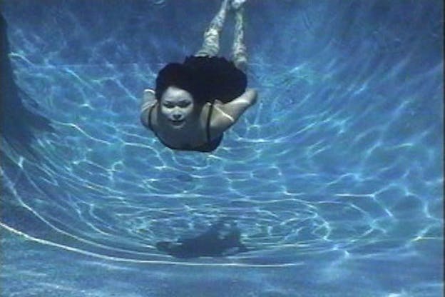 A figure with long dark hair swimming underneath the water in a swimming pool. The sun illuminates creating thin white lines in the bottom of the pool.