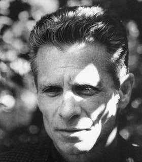 A black and white close up portrait of Michael Gizzi in front of a blurred, spotted background. Dappled light shines on the right side of his face as he looks out into the distance.