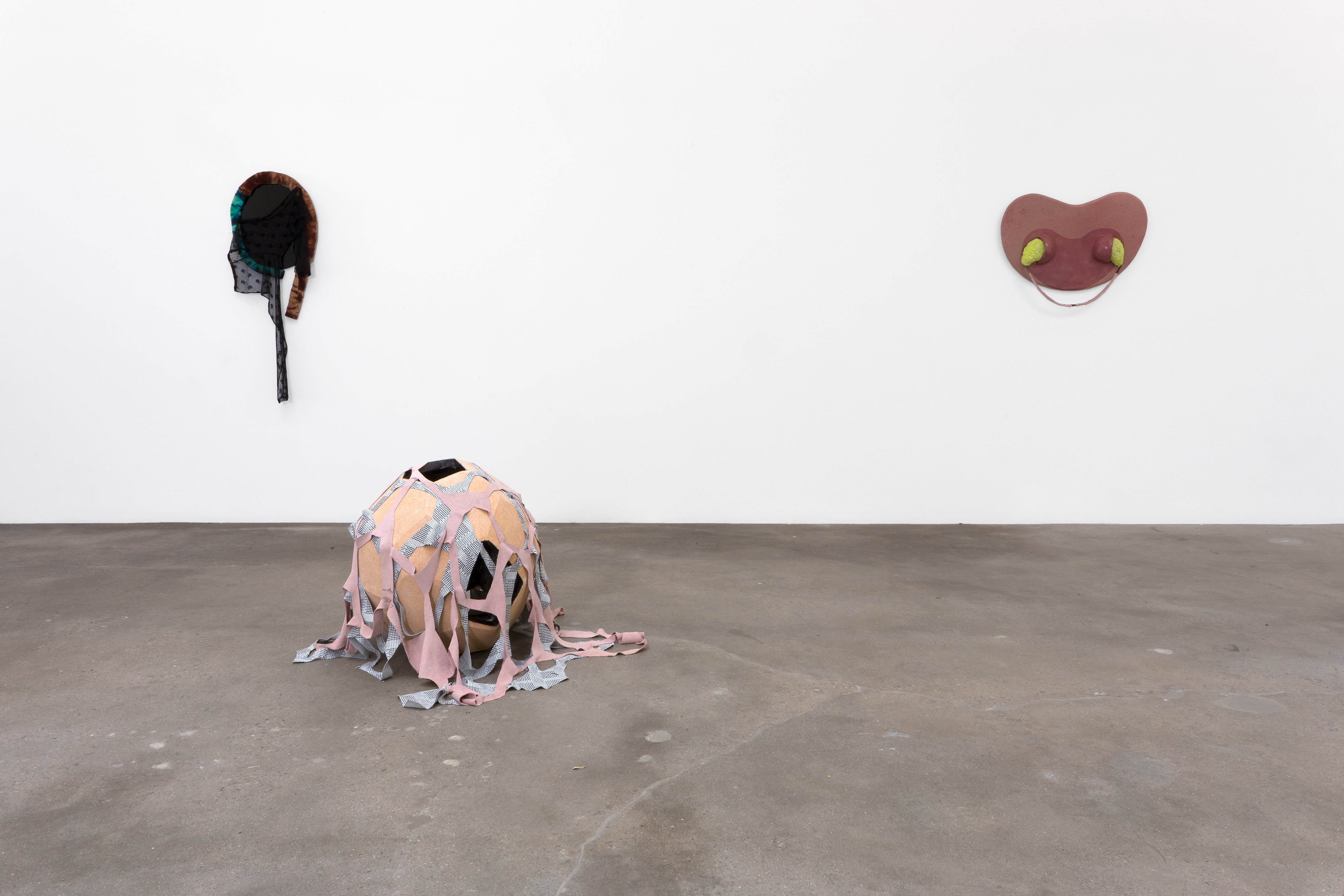 On the left of the image, an ovular, mirror-like sculpture hangs on a white wall with black lacy fabric hanging down from an outer brown and blue velvety coil. On the right, a reddish-pink symmetrically curvilinear sculpture hangs on the wall with neon yellow shapes oozing out on either side. In front of them on the cement floor sits an orb-shaped sculpture draped with fabric netting.