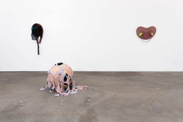 On the left of the image, an ovular, mirror-like sculpture hangs on a white wall with black lacy fabric hanging down from an outer brown and blue velvety coil. On the right, a reddish-pink symmetrically curvilinear sculpture hangs on the wall with neon yellow shapes oozing out on either side. In front of them on the cement floor sits an orb-shaped sculpture draped with fabric netting.
