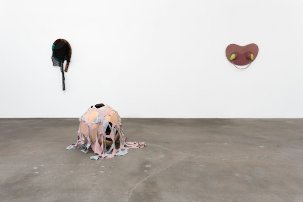 On the left of the image, an ovular, mirror-like sculpture hangs on a white wall with black lacy fabric hanging down from an outer brown and blue velvety coil. On the right, a reddish-pink symmetrically curvilinear sculpture hangs on the wall with neon yellow shapes oozing out on either side. In front of them on the cement floor sits an orb-shaped sculpture draped with fabric netting.