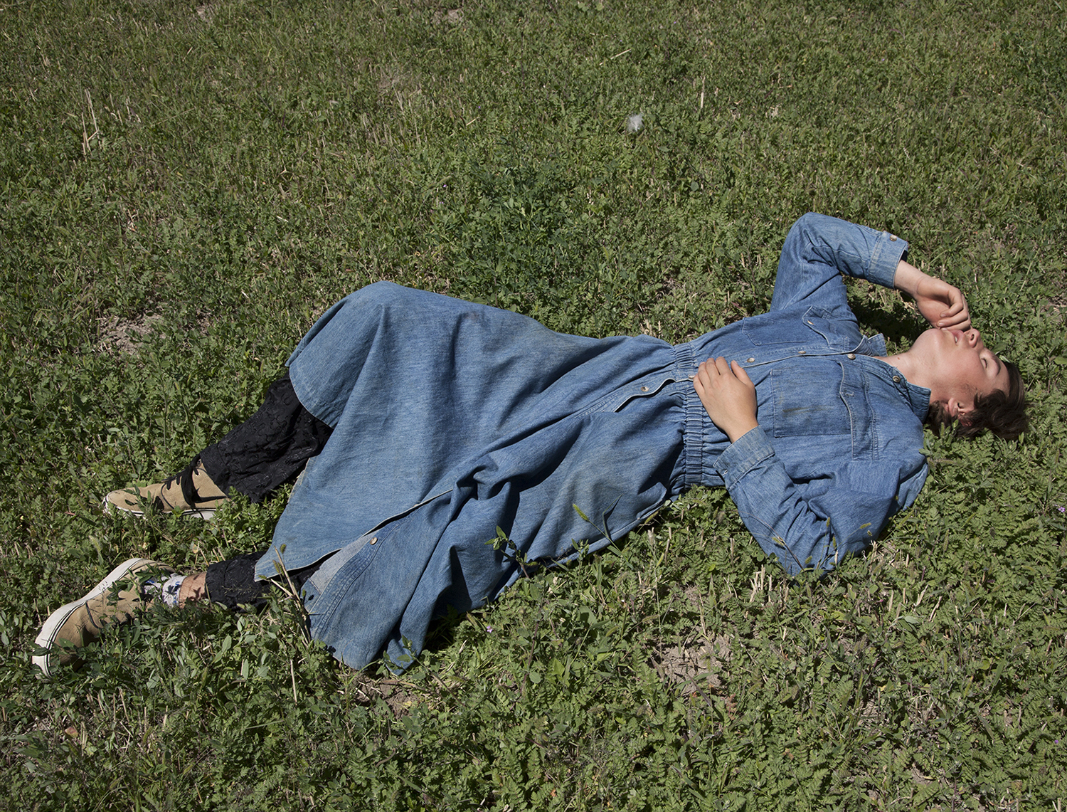 A photograph of a person laying down in grass on a sunny day. They are wearing a long sleeved denim dress that reaches below their knees, black pants underneath, and tan sneakers. Their eyes are closed as one hand reaches up to touch their own cheek, the other rests on their abdomen. Their right leg is bent with their foot flat on the ground and their knee touching the ground.