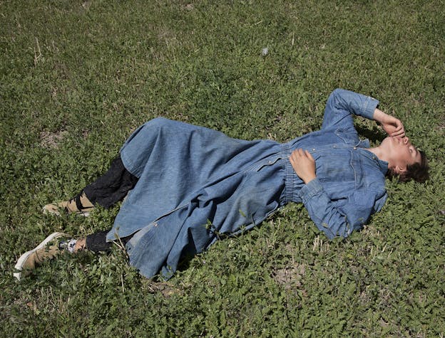 A photograph of a person laying down in grass on a sunny day. They are wearing a long sleeved denim dress that reaches below their knees, black pants underneath, and tan sneakers. Their eyes are closed as one hand reaches up to touch their own cheek, the other rests on their abdomen. Their right leg is bent with their foot flat on the ground and their knee touching the ground.