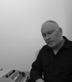 Black and white photograph of a Sean Meehan dressed in a black button up, sitting next to a typewriter and angling his face down towards it.
