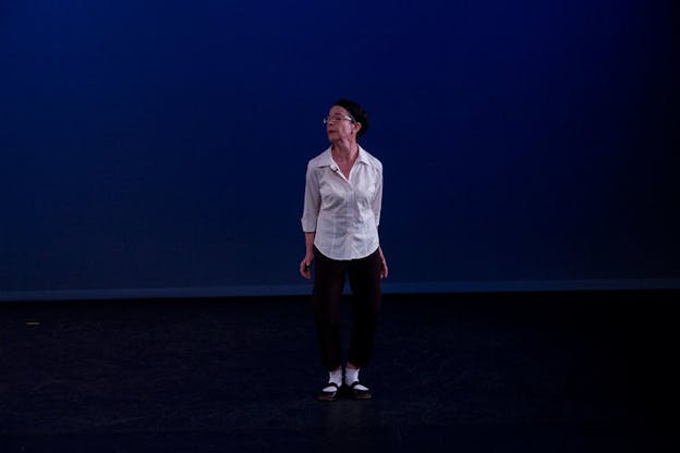 Performer dressed in a white shirt and black pants stands and looks to their right on a dimly lit stage with a black floor and twilight blue backdrop. 