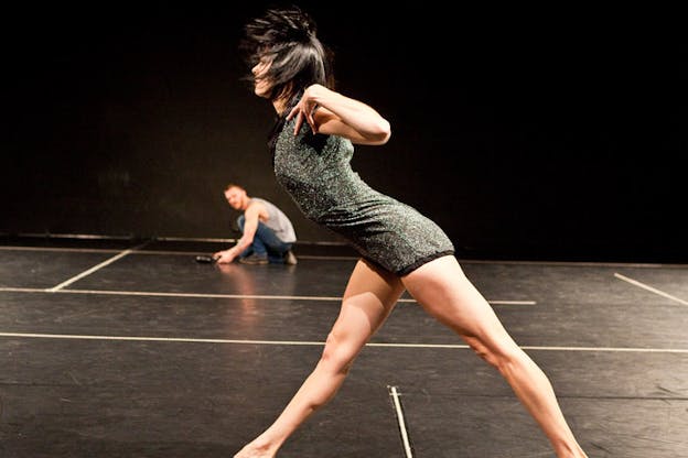 A performer leans forward with their legs spread out. Their arms are lifted with their elbows by their shoulders. They stare intently ahead. They have black hair that is briefly flown into the air around them and they wear a green and black sparkly leotard. In the background, a blurred figure is seen crouching and adjusting something on the floor.  