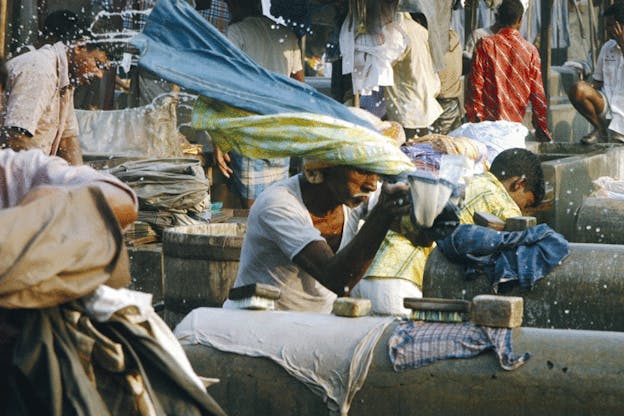 An image of people washing clothes on stone cylinders. The man in the foreground flips a collection of clothing behind him so that water drops splash around him. 