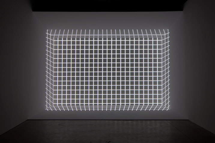 At the far wall of a grey room, a grid made of light is projected in such a way that creates the optical illusion of the grid being a 3D cube.