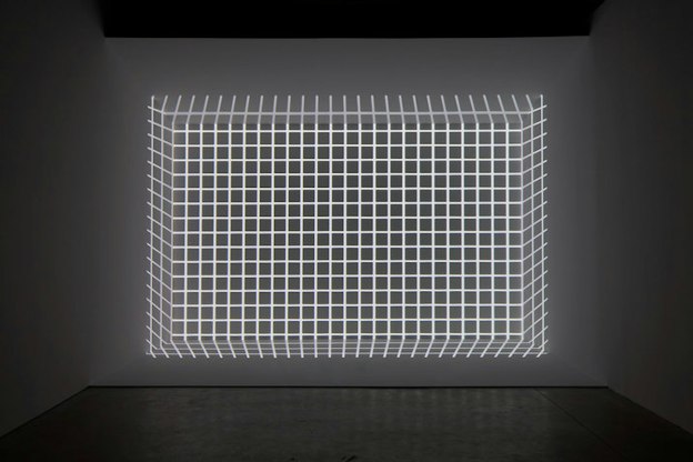 At the far wall of a grey room, a grid made of light is projected in such a way that creates the optical illusion of the grid being a 3D cube.