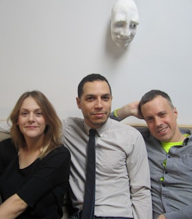 Portrait of My Barbarian in front of a white wall with a white mask hanging from it. On the left, Jade Gordon sits, wearing a black shirt. She has medium length light brown hair. In the middle, Malik Gaines sits, wearing a white button up and a black tie. He has short dark brown hair. On the far right is Alexandro Segade who has short brown hair and is wearing a light grey button up over a neon green shirt. 