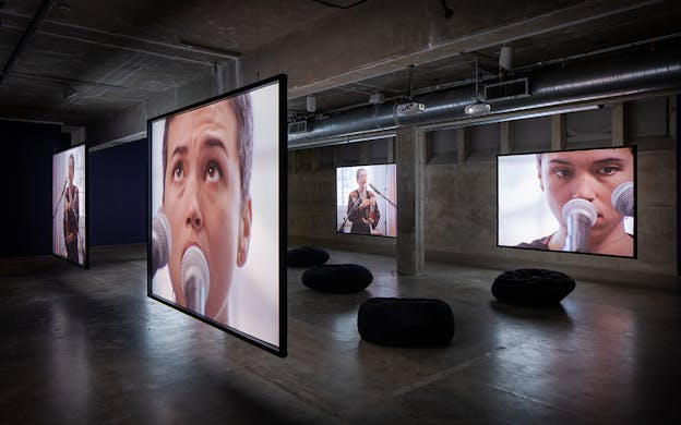 Four screens are hanging in a square, illuminated with images of Holland Andrews in various stages of performance. There are black bean bag seats on the ground in between the screens.