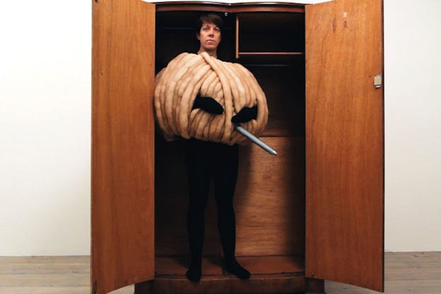 A person stands in a wooden dresser dressed in a black unitard and around their torso and chest a huge ball of yarn with an oversized needle sticking through. 