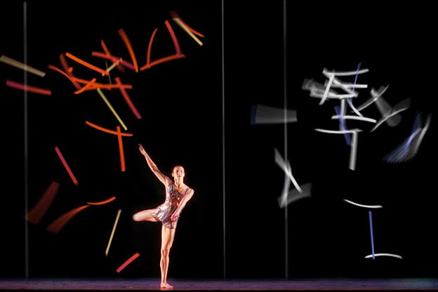 One performer dances in the lower left of a large dark stage. They wear a metallic leotard. Resting only on the left leg, they lift both arms up and hold their right leg out behind them. Projections of scattered lines fill the back wall. On the left, the lines are mostly red with a few gold and white ones and on the right, the lines are mostly white with three blue ones scattered in. 