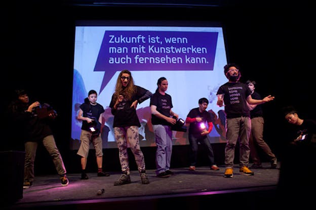 Performers wearing matching black tee-shirts –one performer wearing groucho glasses–hold lights and make gestures on a stage with screen projecting a dialogue box in german.