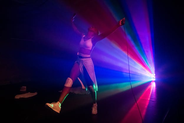 A person dressed in white enthusiastically raises their arms up and walks as a light with colors of red, purple, green and blue lights them from behind.