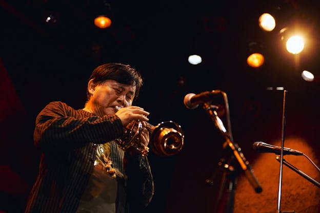 Koichi Makigami is photographed from below as he performs on a stage with orange toned lighting. He stands on the left side of the frame playing a trumpet with a mute. He is wearing a dark striped buttoned shirt over a t-shirt. 