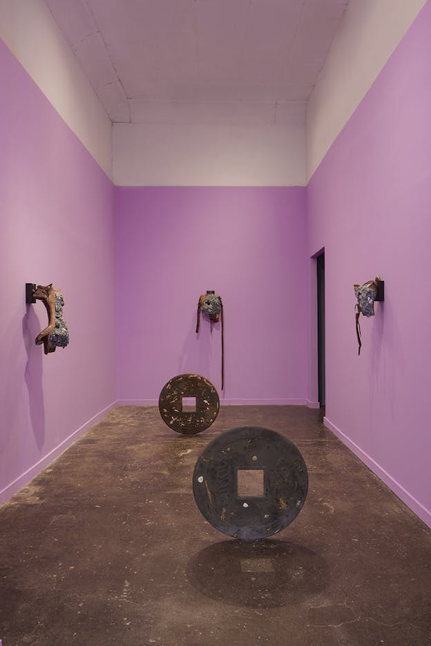 Five sculptures installed in a gallery with pink walls and a cement floor. Three sculptures are mounted on each of the three visible walls, and two others, circular shapes standing upright with squares cut out of the middle, are on the floor.