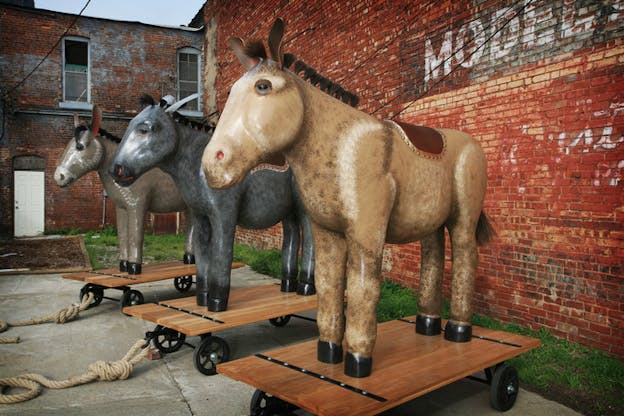 Three donkeys on dollies stand in front of a red brick building. The donkey on the far left is light grey, the middle one is a darker grey and has a ribbon on its head, and the donkey on the far right is tan.
