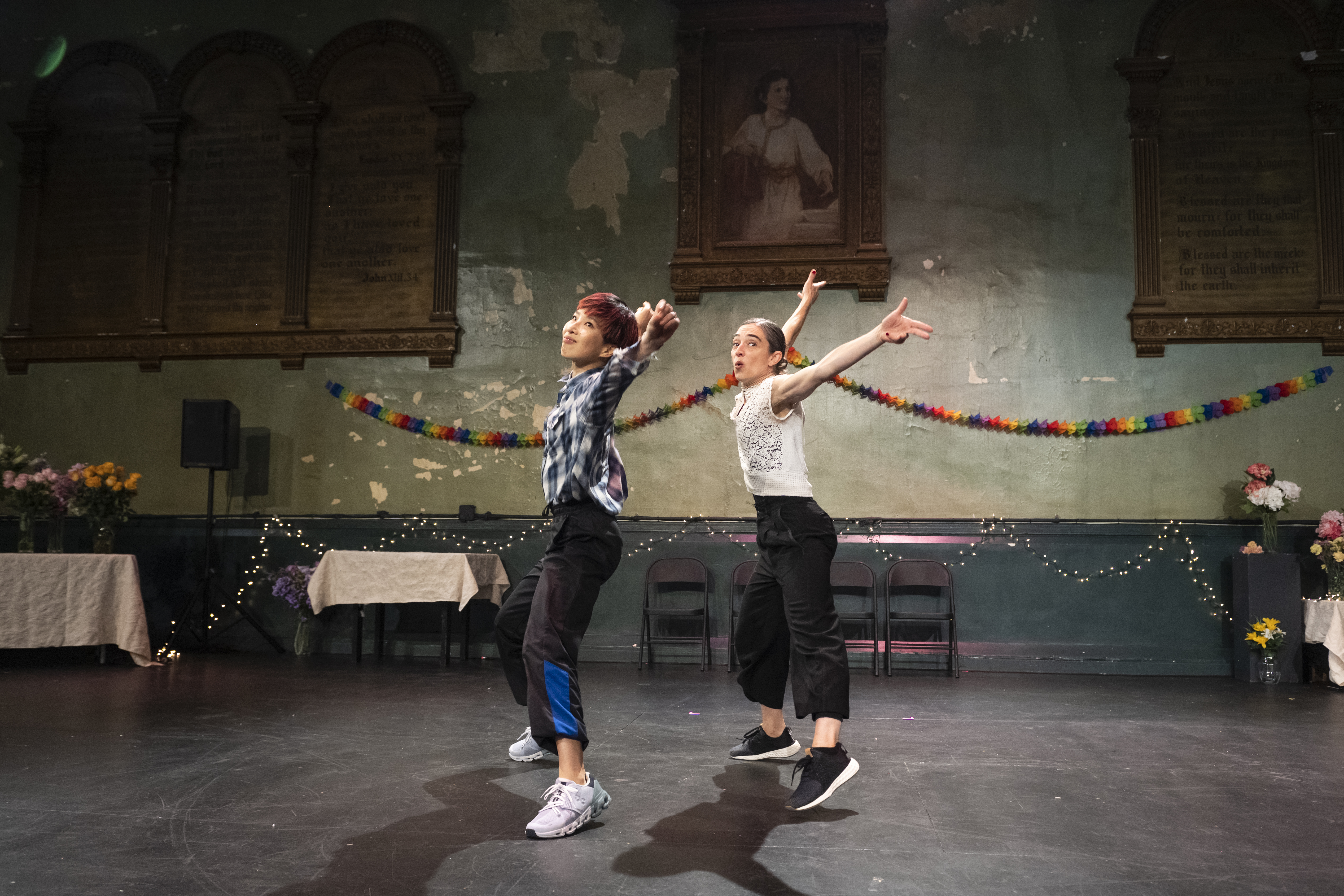 Two performers dance in the middle of a space decorated for a party. The two dancers are centered in the frame, both facing the left and standing in jumping jack positions with their feet wide apart and arms raised, their heads are turned to look at the camera. Hsiao-Jou Tang dances on the left half of the frame with a smile on her face, gazing above the camera and making snaps with her hands. She is wearing a blue and white flannel button up, black sweatpants with blue stripes above the cuffs, and white sneakers. Monica Bill Barnes dances on the right of the frame, also gazing upwards. She is wearing a white sleeveless blouse with a lace front, black high-waisted pants, and black sneakers.